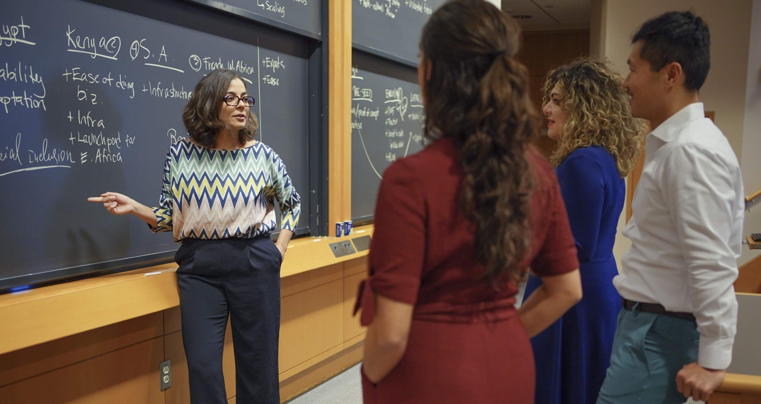Paula stands in front of a blackboard, engaged in a discussion with three others