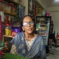 Woman sitting inside her convenience store, surrounded by shelves of snacks and a refrigerator of beverages.
