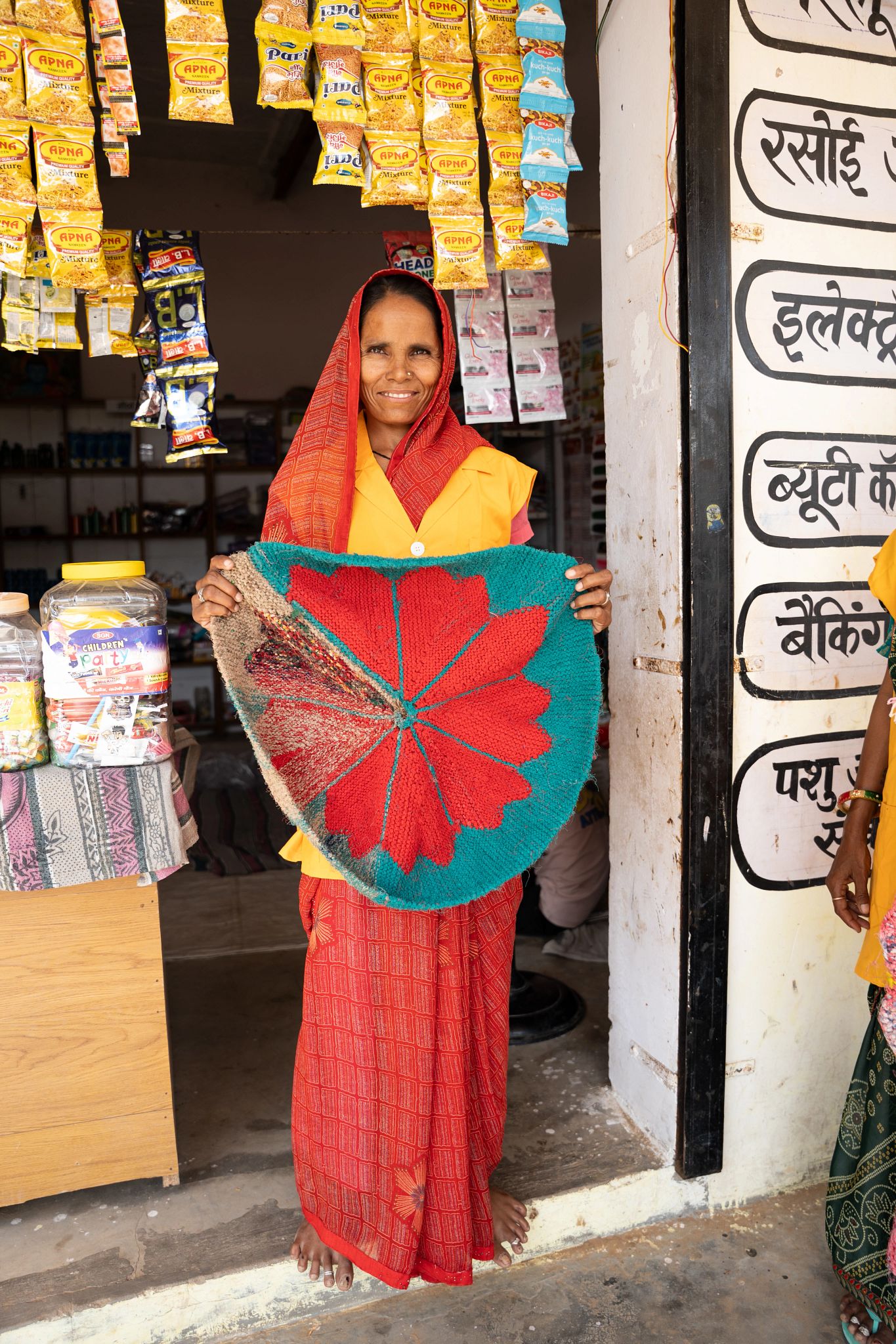 A rural woman entrepreneur standing outside her village shop in Rajasthan, smiling and holding a handwoven carpet she sells as part of her business.