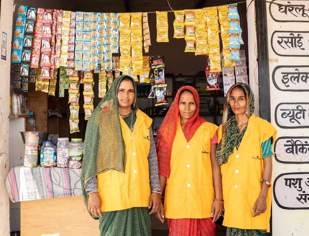 Three women wearing yellow vests stand outside a village shop named Savitri Saheli Super-Mall in Rajasthan.