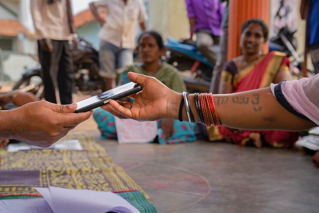A woman hands a mobile phone to another person during a financial transaction in a community setting.